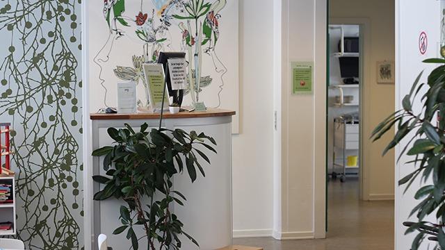 Moderne reception med planter og vægmaleri, der fører ind til et behandlingsrum.