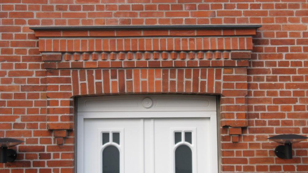 Detalje af en murstensvæg med en dekorativ overligger over en hvid dør med buede vinduer.