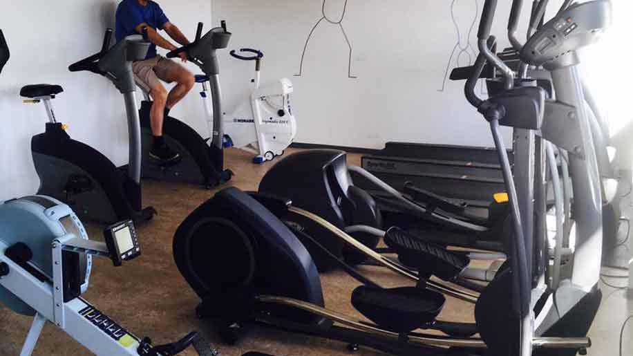 En person træner på en motionscykel i et fitnesscenter med forskellige træningsmaskiner i baggrunden.