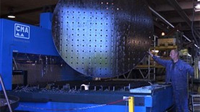 En mand arbejder med en stor perforeret metalplade på en industriel maskine i en fabrik.