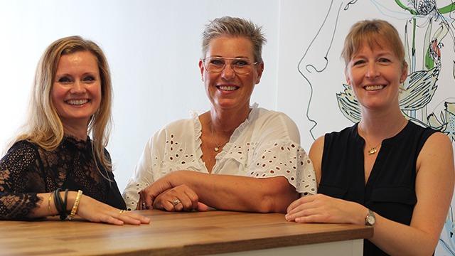 Tre kvinder smiler og står ved en trædisk i et lyst rum med grønne planter i baggrunden.