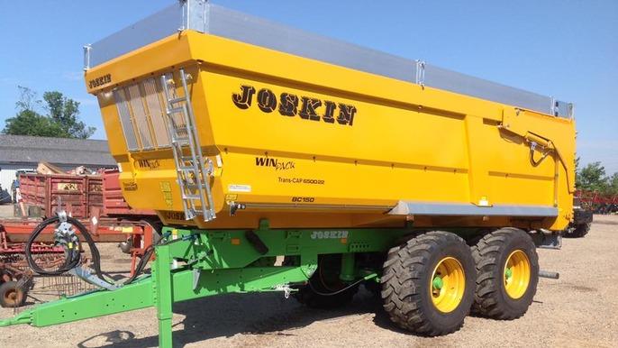 Gul Joskin vogn med store hjul og grøn chassis, parkeret på en gård under en klar blå himmel.