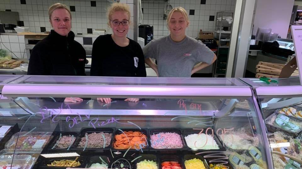 Tre kvinder står bag en disk med forskellige pålæg og toppings i en delikatesseforretning.