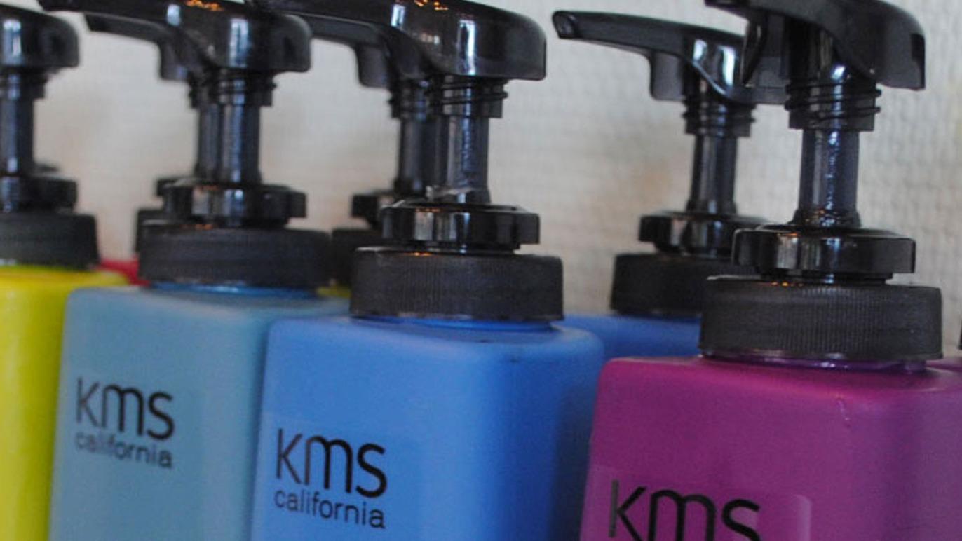Farverige KMS California shampoo- og balsamflasker med pumpe på en hylde.