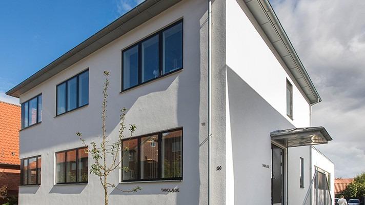 Moderne hvidt hus med store vinduer og en indgang under et tag. Grønt træ foran bygningen.