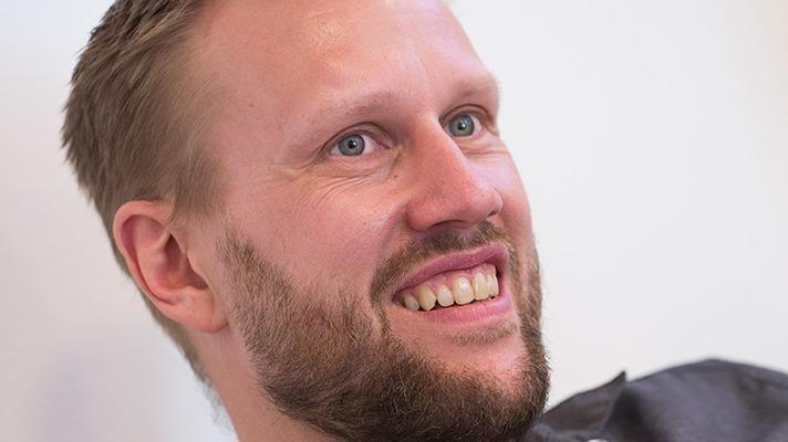 En mand med kort hår og skæg smiler, mens han ser op, med et venligt udtryk i ansigtet.