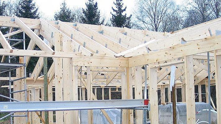 Byggeri af et hus med træramme og tagkonstruktion, der viser stilladser og materialer i en skovagtig omgivelser.