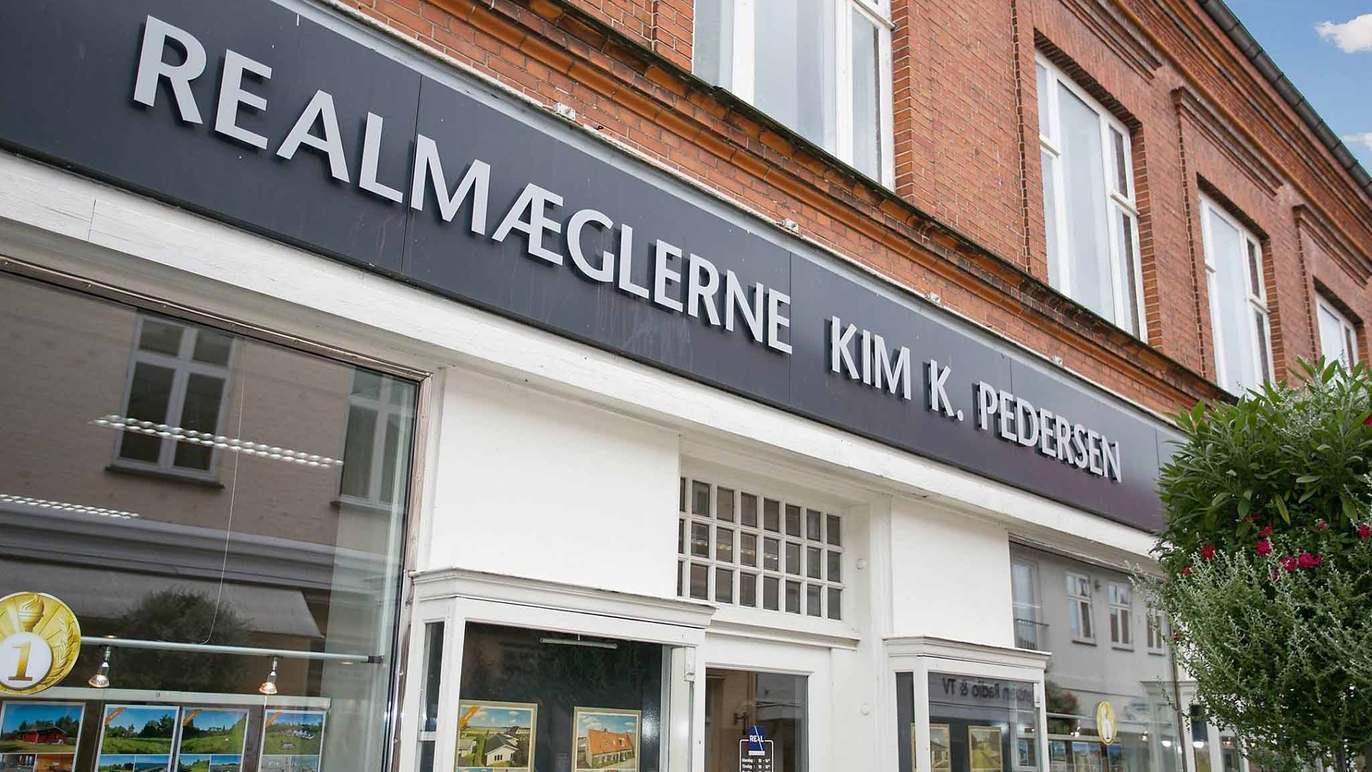 Ejendomsmæglerbutik med skiltet "RealMæglerne Kim K. Pedersen" på en murstensvæg.
