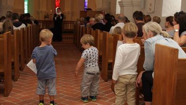 Tre børn står i en kirke og ser på en præst, mens voksne sidder på bænke i baggrunden. Farverigt lys fra vinduerne.