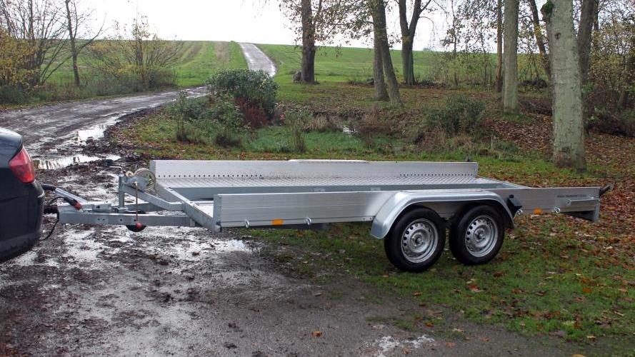 En sølvfarvet trailer til bil, parkeret på en mudret vej omgivet af træer og græs.