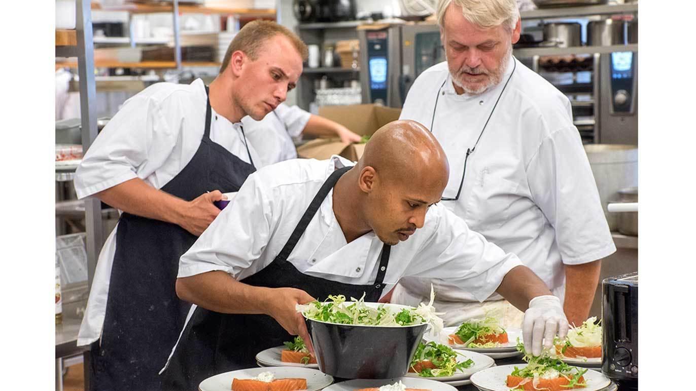 Tre kokke arbejder sammen i et køkken, mens de anretter mad på tallerkener med salat og grøntsager.