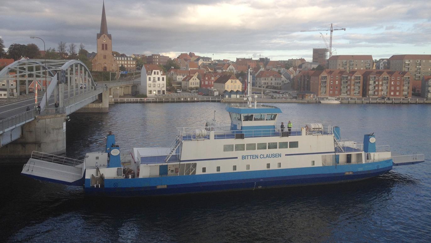 Færgen "Cittylink" ligger ved kajen med en by i baggrunden og en bro, der forbinder de to sider.