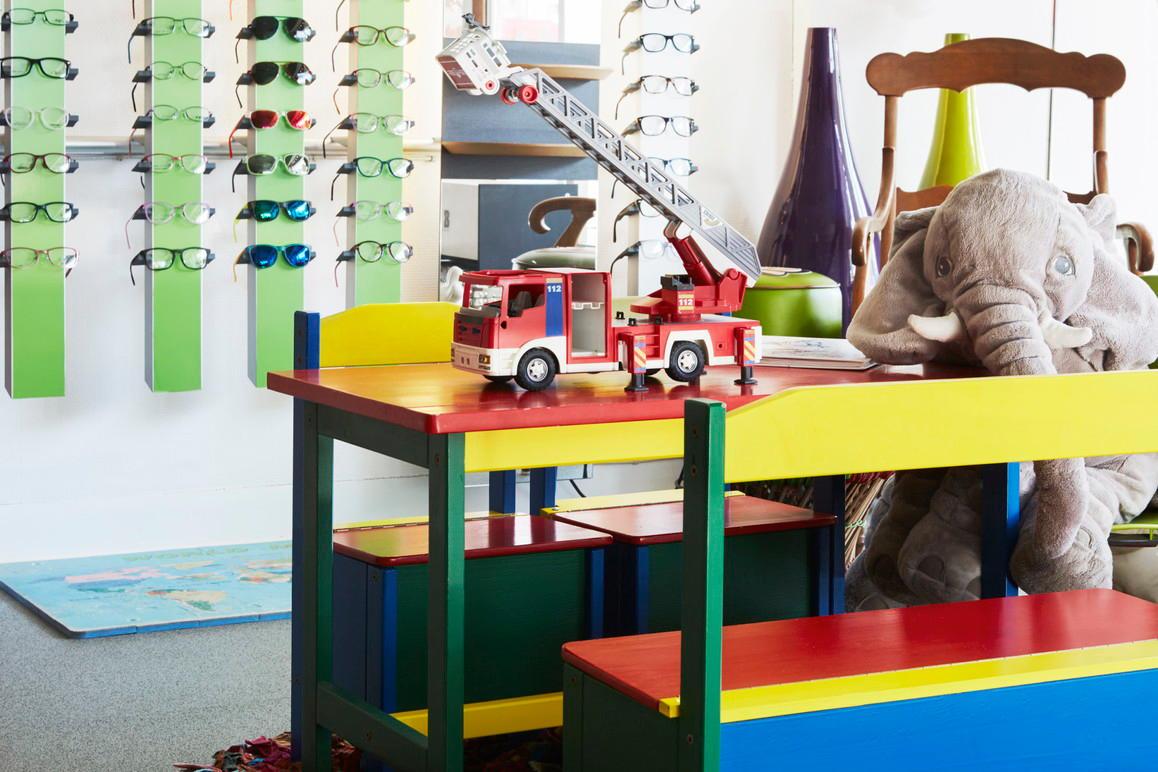 Farverig legeplads med legetøjsbrandbil, elefant og briller på væggen i en børnevenlig butik.