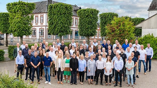 En gruppe på 50 personer står samlet foran en bygning med grønne træer og en blå himmel i baggrunden.