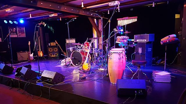 Scene med musikudstyr på en scene, herunder trommesæt, guitarer og percussion-instrumenter.