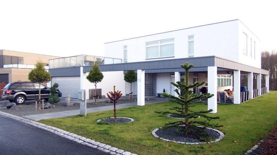 Moderne hus med fladt tag, grøn græsplæne og parkerede biler foran. Træer og buske pryder indgangen.