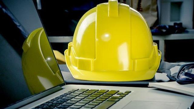 Gul sikkerhedshjelm placeret på en bærbar computer i et arbejdsområde med værktøj i baggrunden.
