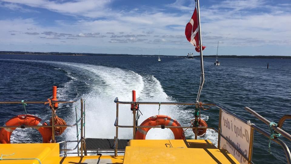Udsigt fra en gul færge med redningskranse, der sejler på blåt vand med bølger og en dansk flag.