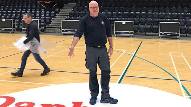 En mand står på en basketballbane, mens en anden person arbejder i baggrunden med at rydde op.