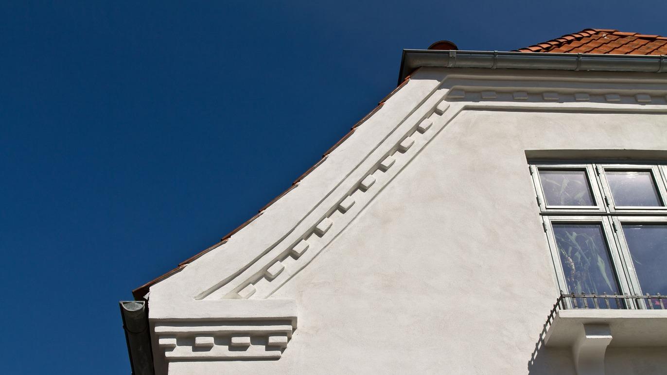 Husets hjørne med hvid facade og detaljeret gesims mod en klar blå himmel.