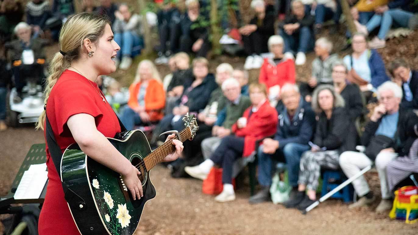En kvinde i rød kjole synger og spiller guitar for et publikum i en skov. Publikum sidder på bænke og lytter.