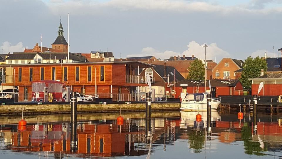 Havn med træbygninger og både, der spejler sig i vandet under en klar himmel.