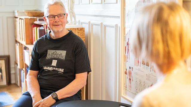 Mand i sort t-shirt sidder ved et bord og taler med en kvinde i et lyst rum med bøger og billeder.