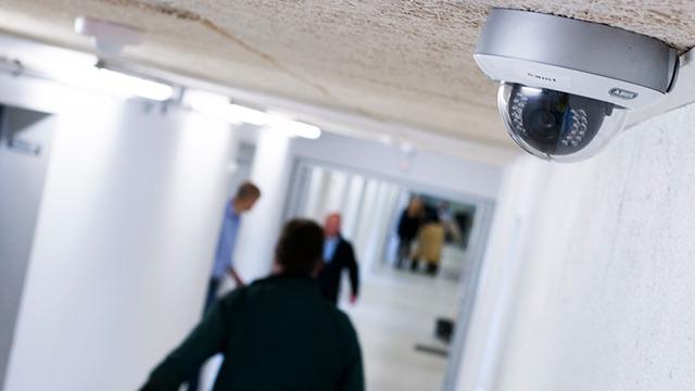 Overvågningskamera i en gang med personer i baggrunden, der går mod en dør.