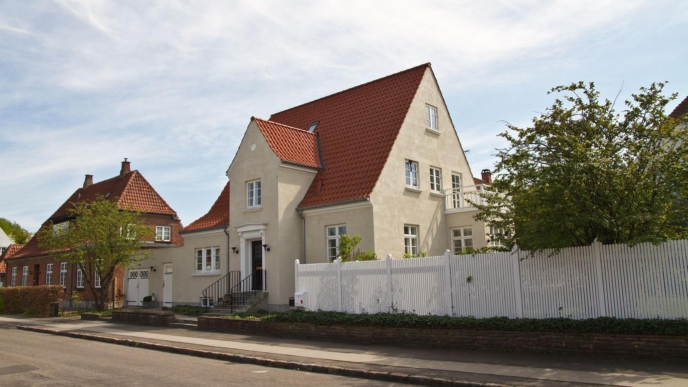 En hvid villa med rød tag og hvidt hegn, beliggende på en stille gade med træer i baggrunden.