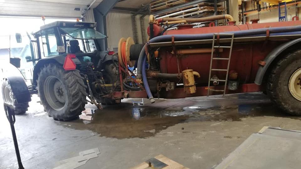 Traktor med gyllevogn i en garage, omgivet af værktøj og vandpytter på gulvet.