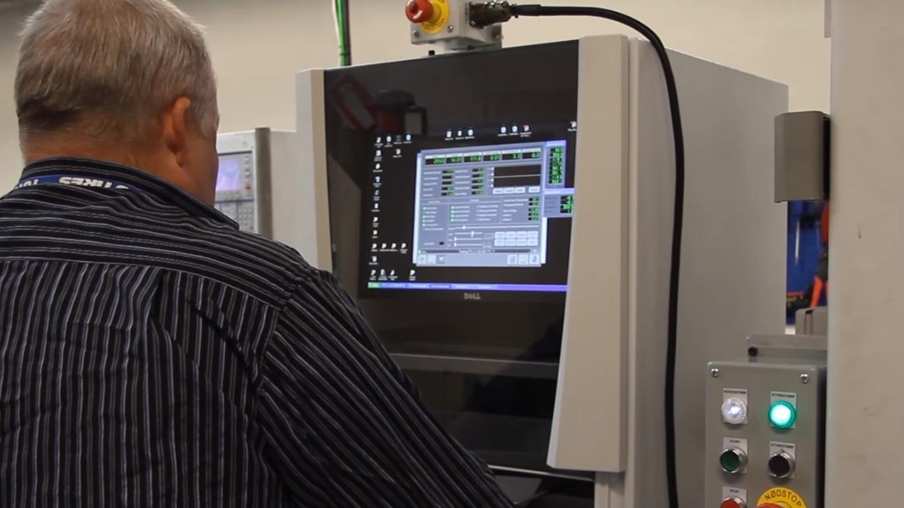 En mand arbejder ved en maskine med en digital skærm og kontrolpanel i en produktionshal.