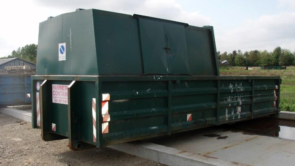 Grøn affaldscontainer placeret på en grusvej med græs og træer i baggrunden.