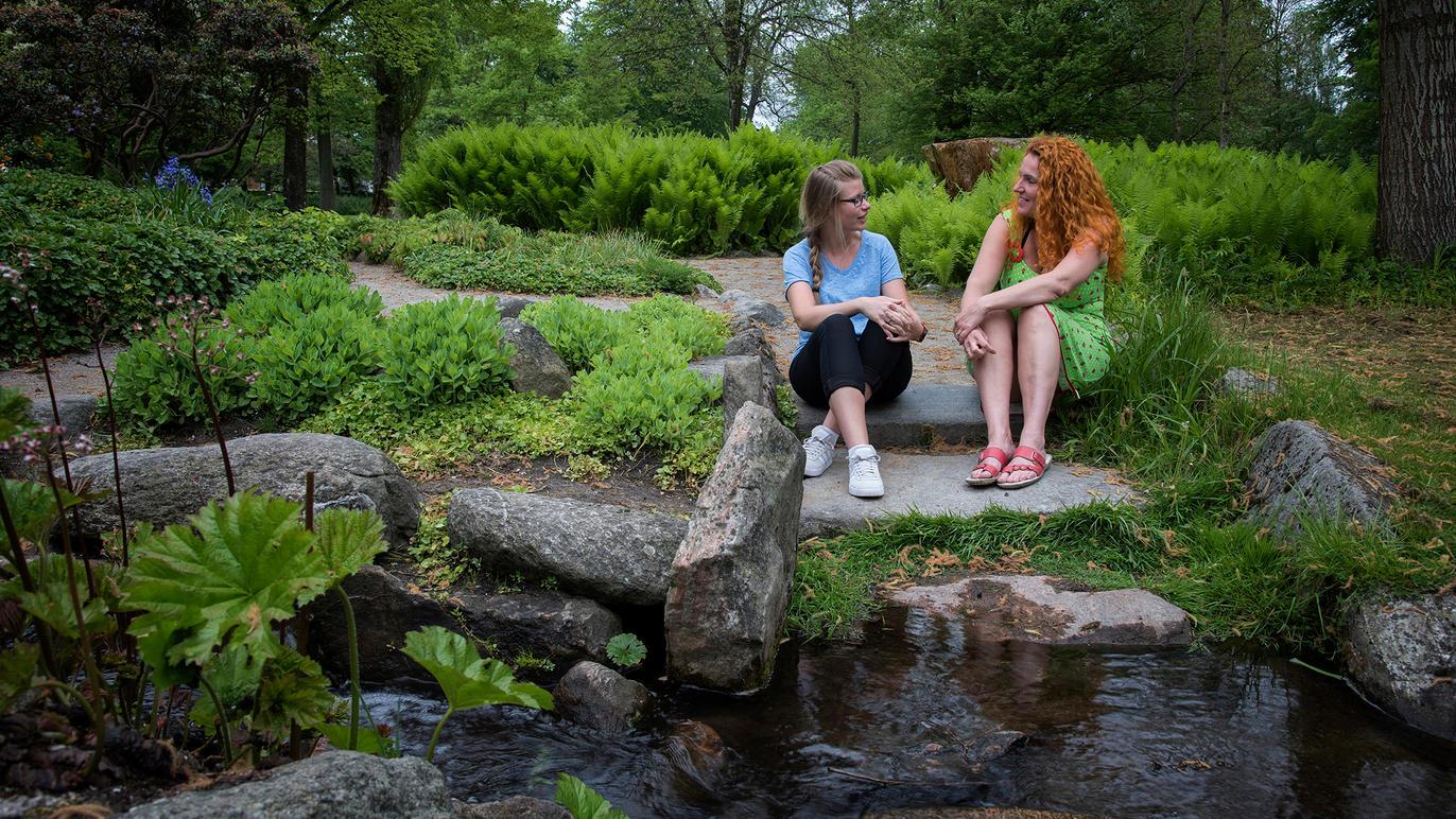 To personer sidder på en sten ved en lille bæk i en grøn park med buske og træer omkring.