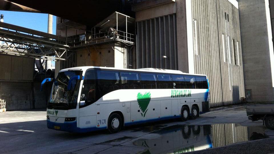 En bus parkeret ved en bygning med industrielle strukturer og reflektioner i vandet.