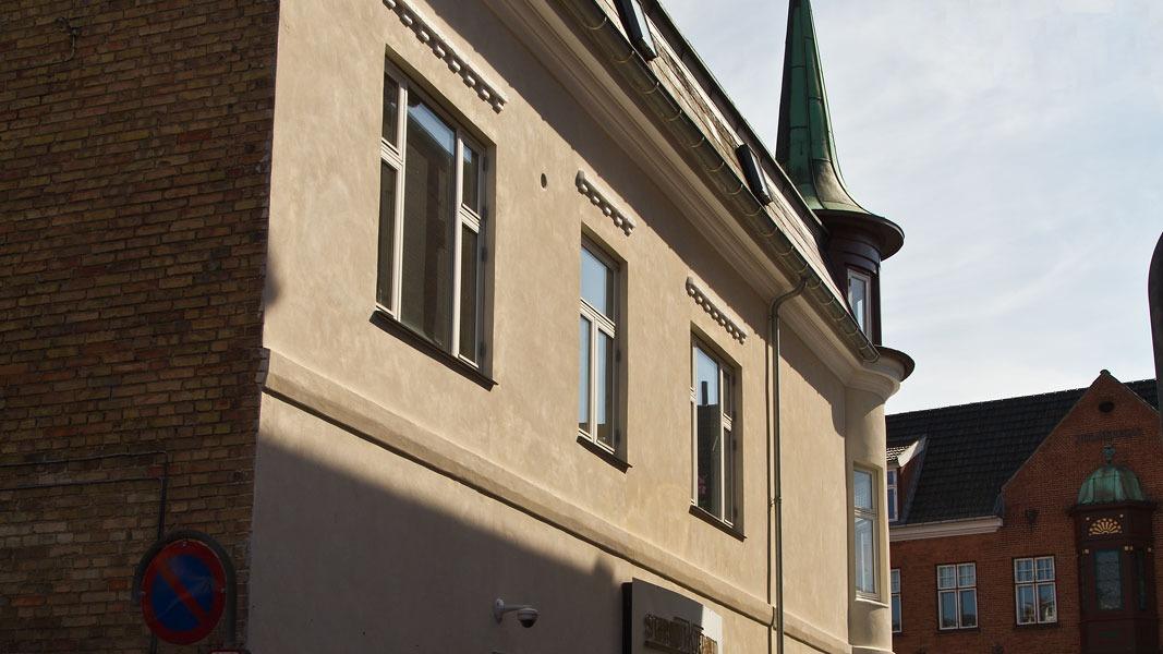 En bygning med en spids grøn tag og vinduer, beliggende på en gade med sollys og skygger.