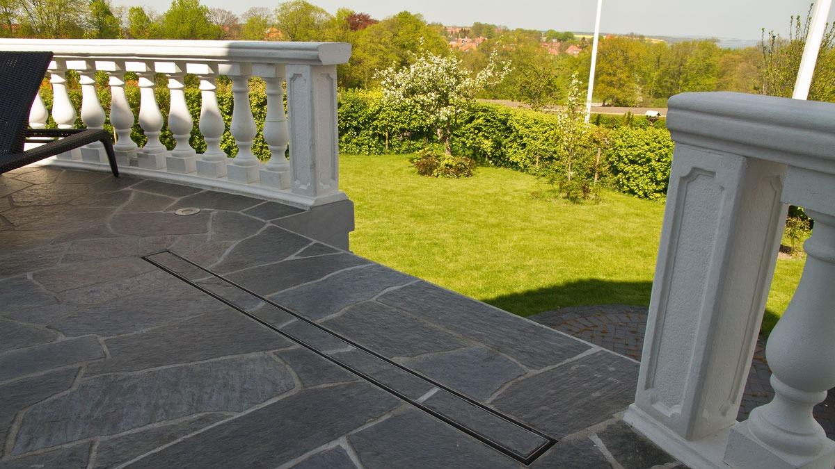 Terrasse med grå stenbelægning, hvid rækværk og grøn græsplæne i baggrunden.