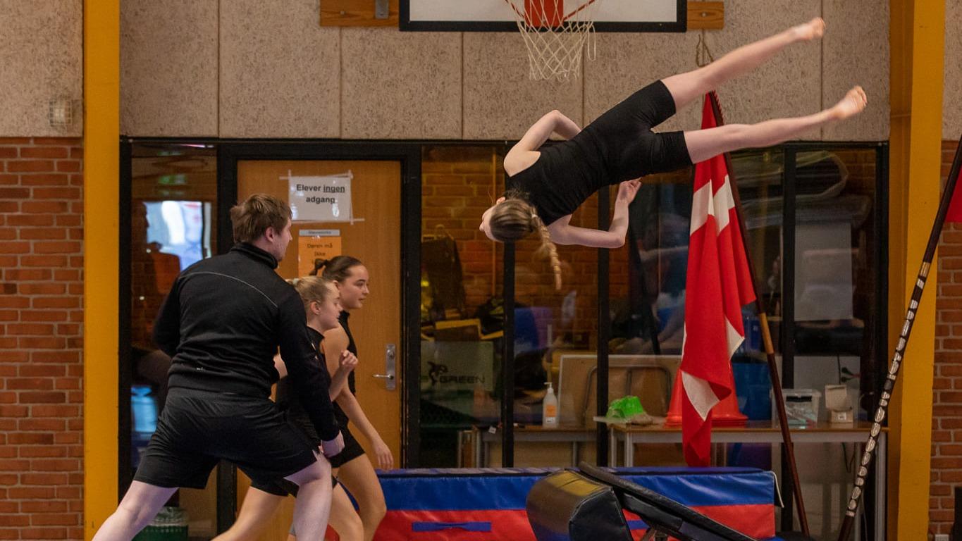 Gymnast udfører en salto over en trampolin, mens trænere og andre gymnaster ser på i en hal.