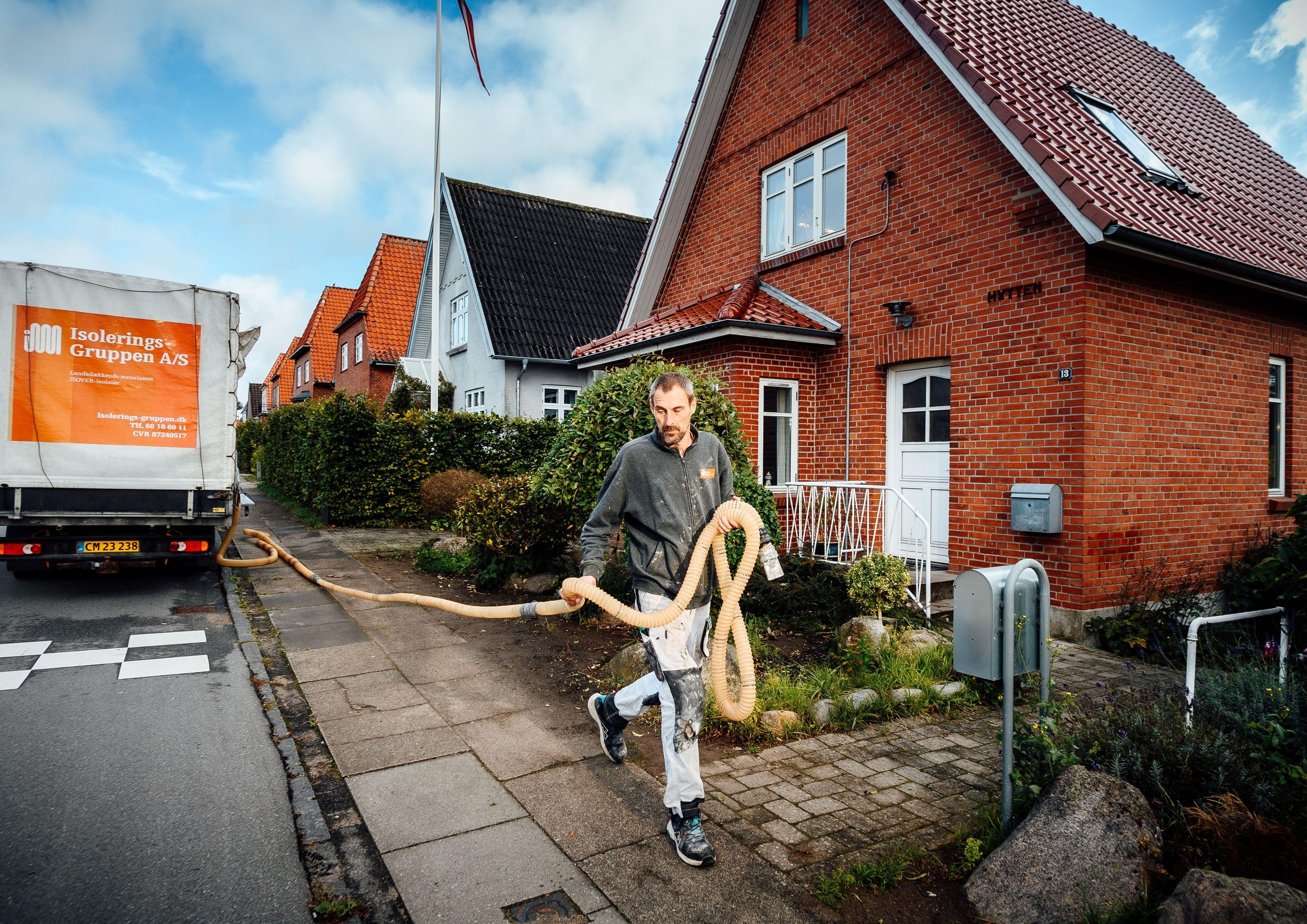 En mand bærer en lang slange foran et rødt hus, mens en flyttebil holder parkeret på gaden.