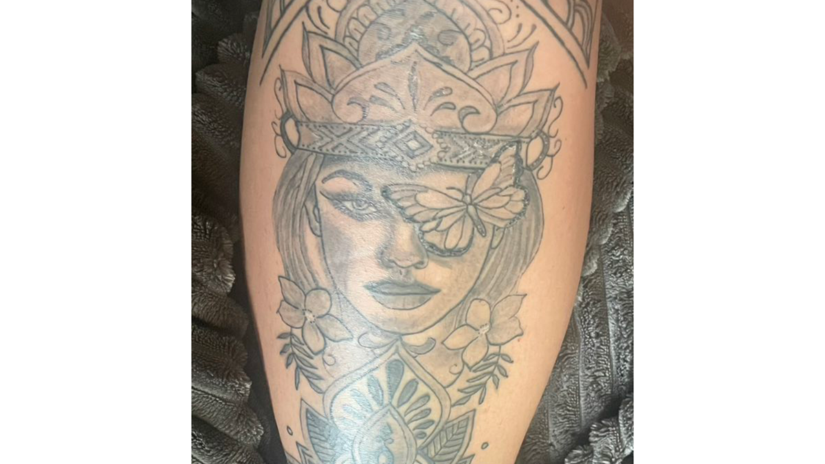 Tatovering af en kvinde med en krone og sommerfugl, dekoreret med blomster på en skinneben.