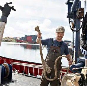 En mand i arbejdstøj holder et reb på en båd ved havnen, med vand og bygninger i baggrunden.