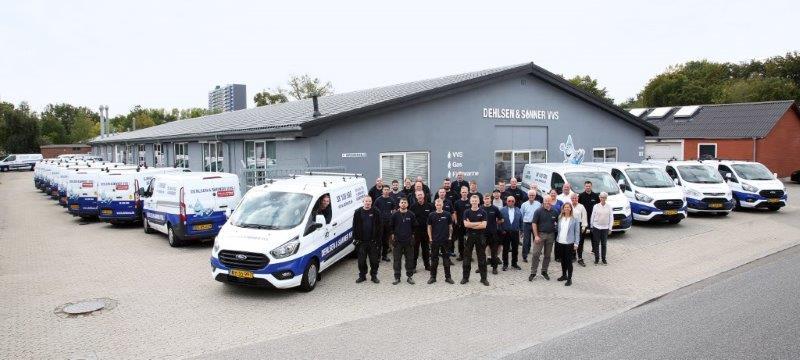 En gruppe medarbejdere står foran en række servicevogne ved en virksomhed i et industriområde.