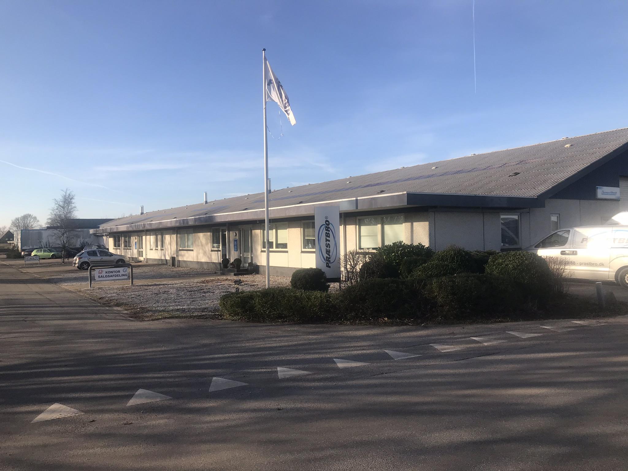En bygning med fladt tag og flag, omgivet af buske og parkerede biler på en solrig dag.