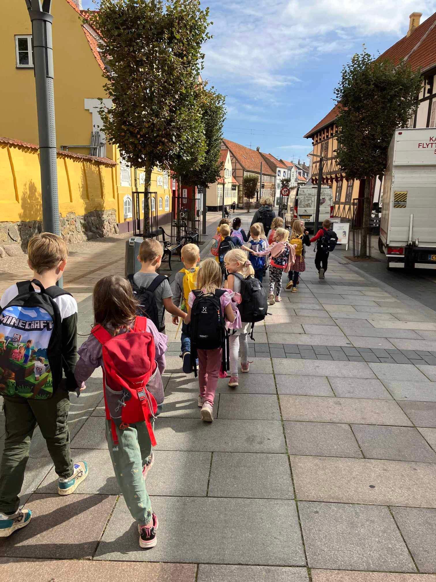 Børn går hånd i hånd ned ad en gade med farverige bygninger og træer langs fortovet.