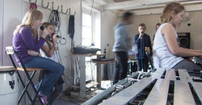Børn synger og spiller musik i et øvelokale med instrumenter og udstyr.