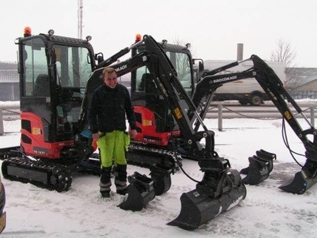 En mand står mellem to gravemaskiner med sne på jorden, klar til at rydde sne.
