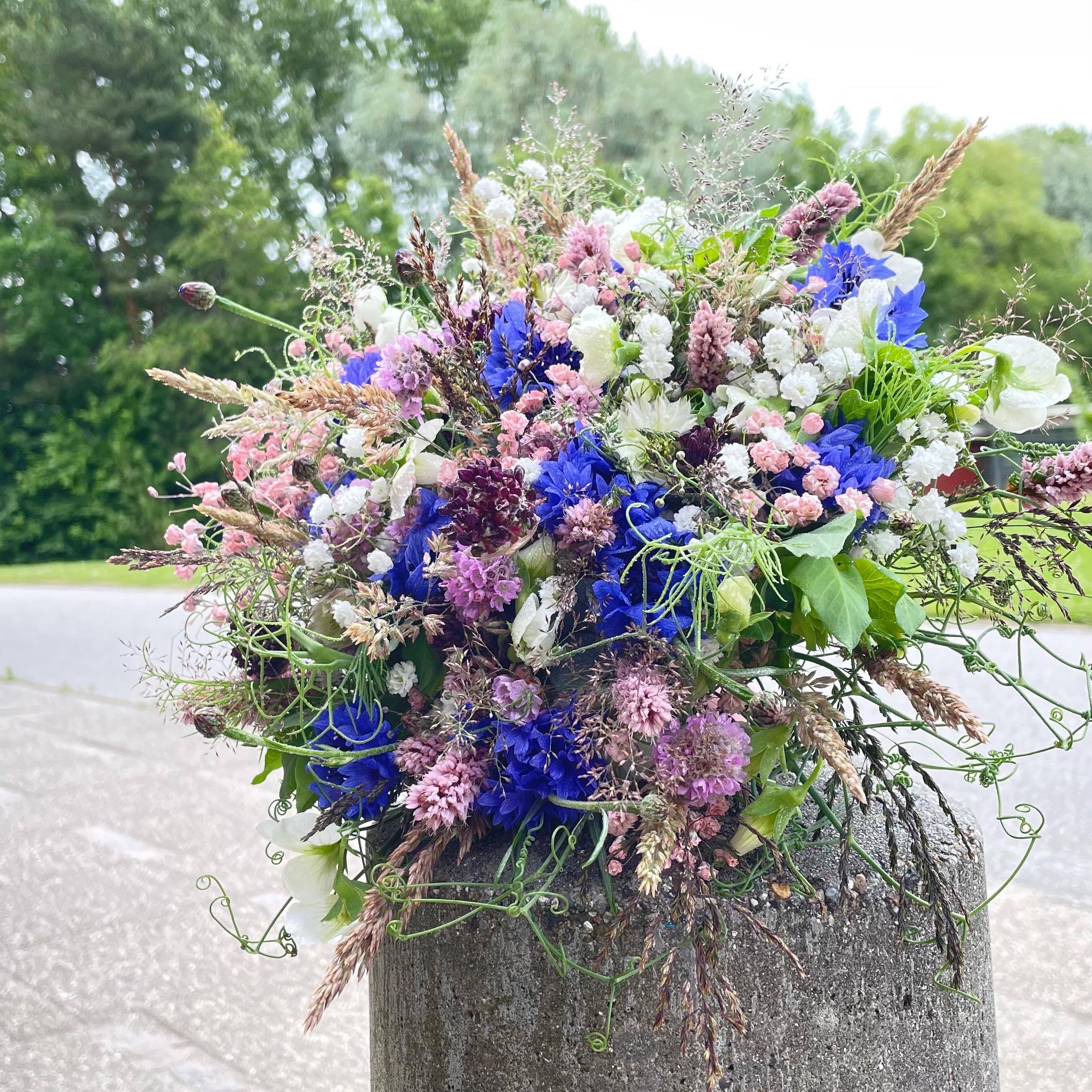 En farverig buket med blå, lilla og lyserøde blomster i en grå krukke ved en vej.