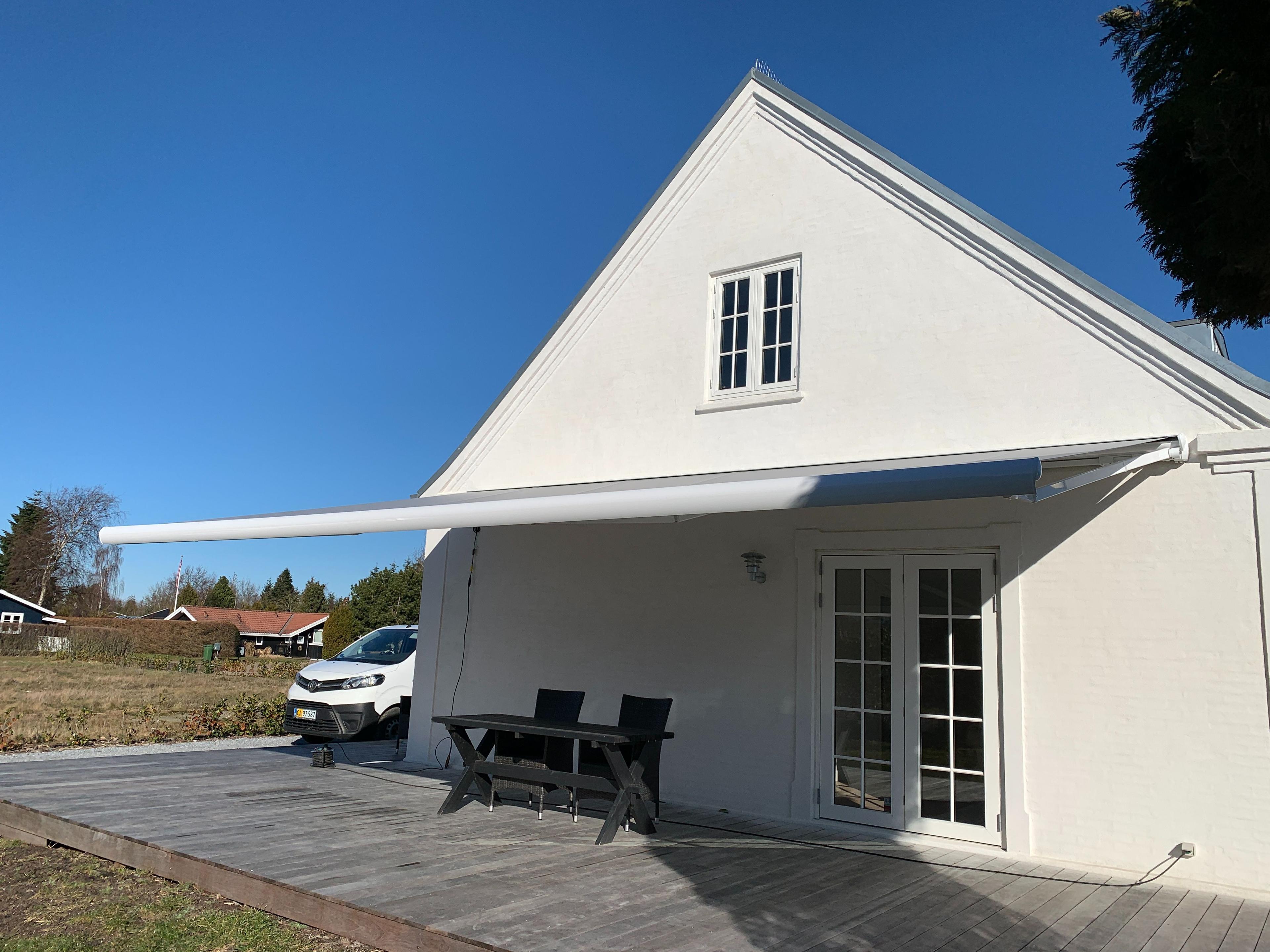 Hus med hvid facade og markise, terrasse med borde og stole, samt en bil parkeret ved siden af.