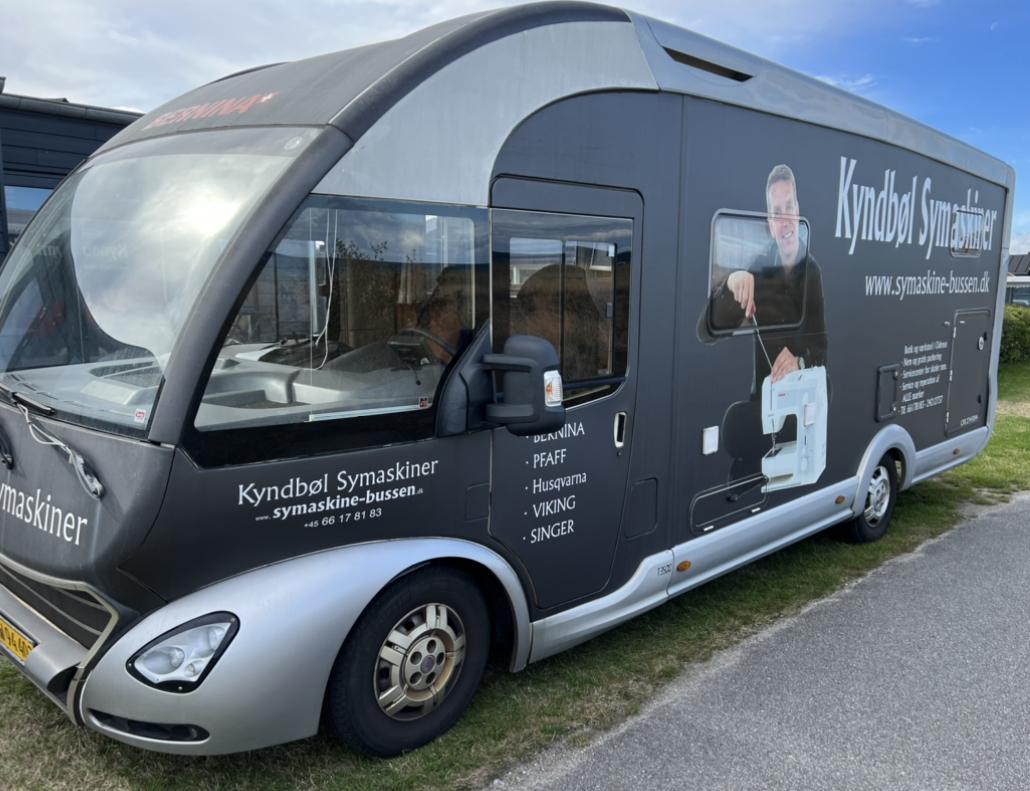 En grå mobil symaskinebutik med tekst og logo på siden, parkeret på en græsplæne.