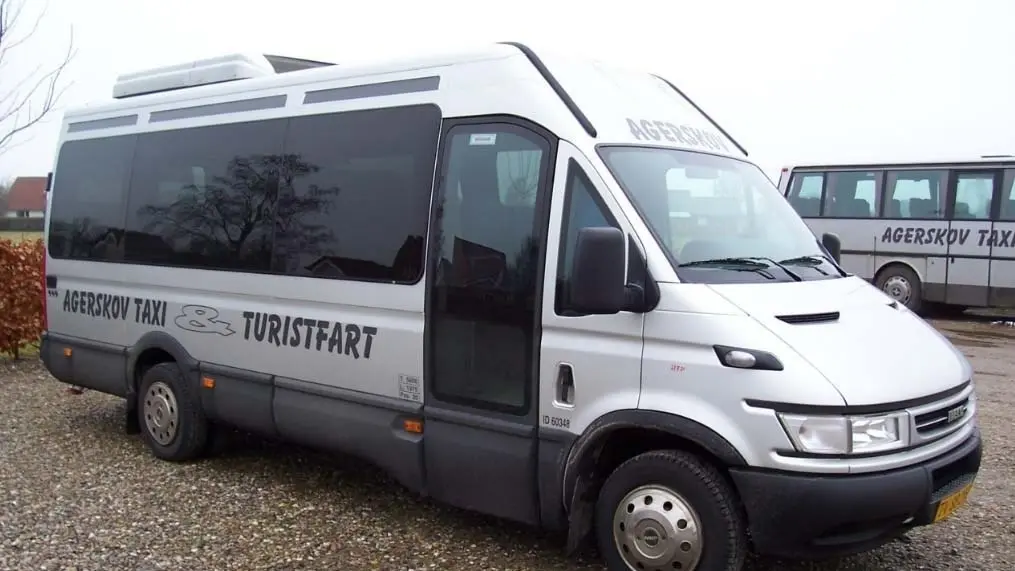 En sølvfarvet minibus med teksten "Afrejse Taxi Turistfart" på siden, parkeret på en grusvej.