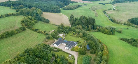 Luftfoto af en stor ejendom omgivet af grønne marker og skove i et naturskønt landskab.
